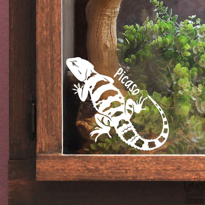 Decorative lizard decal affixed on the glass with 'Picaso' on an enclosure door  with plants and wood.