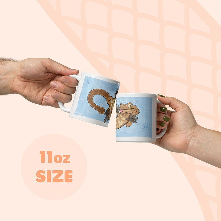Two hands holding white 11oz mugs with blue cartoon crested gecko illustrations, clinking them together against a pale peach background with a subtle snake scale pattern.