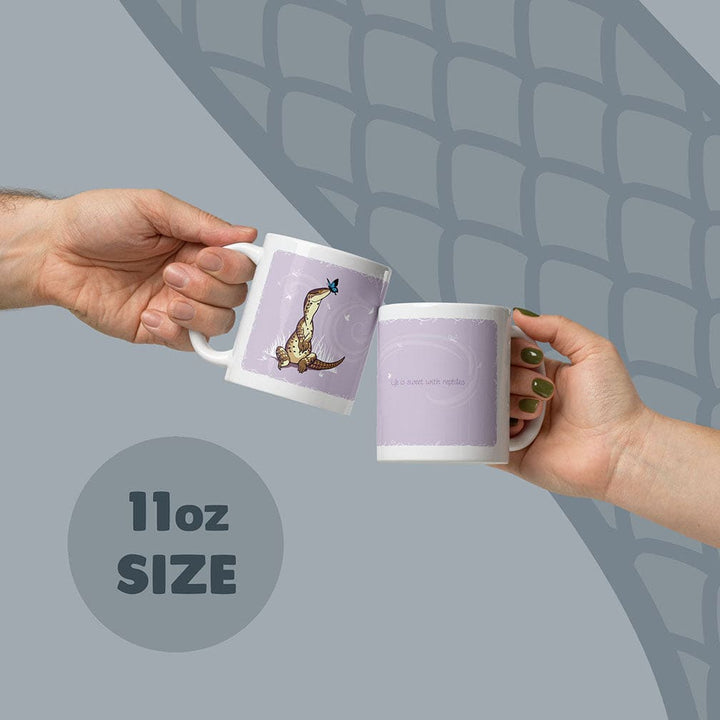 Two white 11oz ceramic mugs with a light purple background. One mug features a brown lizard with a blue butterfly on its nose. The other mug displays the text &