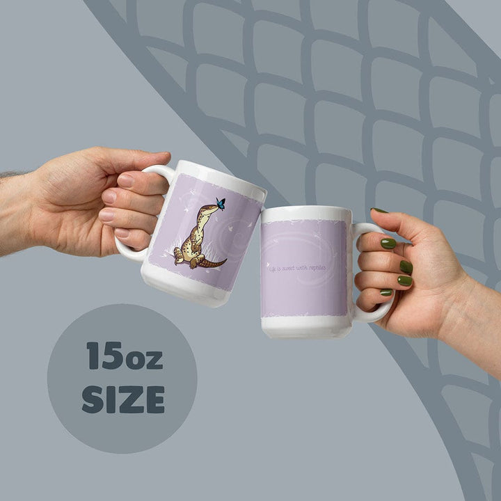 Two white 15oz ceramic mugs with a light purple background. One mug features a brown lizard with a blue butterfly on its nose. The other mug displays the text &