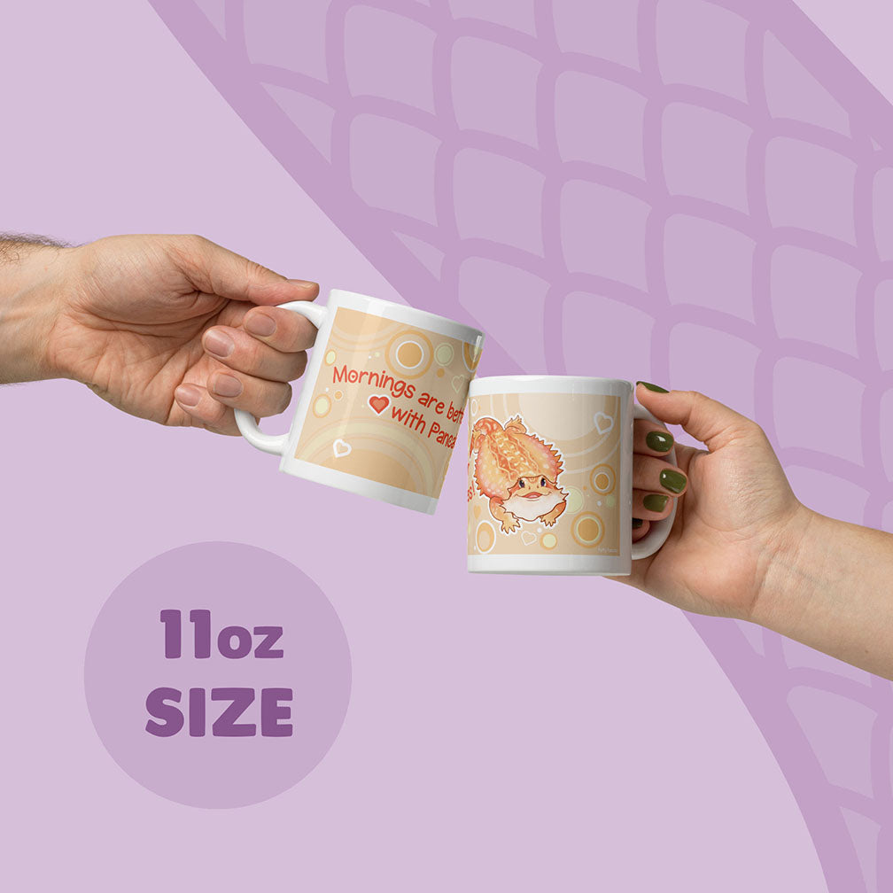 Two hands holding up two 11oz ceramic mugs with cute cartoon bearded dragon and text, set against a light purple background with a subtle scale pattern.
