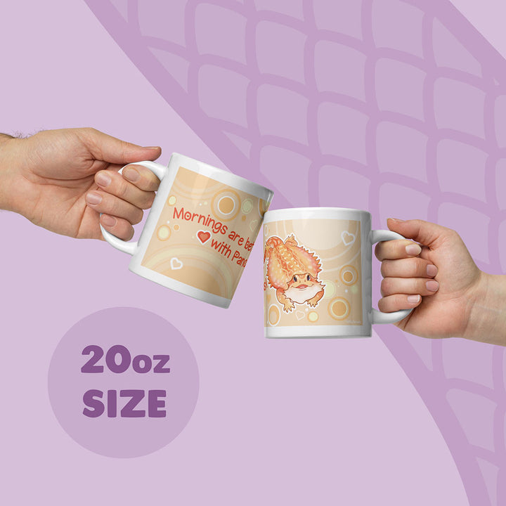 Two hands holding up two 20oz ceramic mugs with cute cartoon bearded dragon and text, set against a light purple background with a subtle scale pattern.
