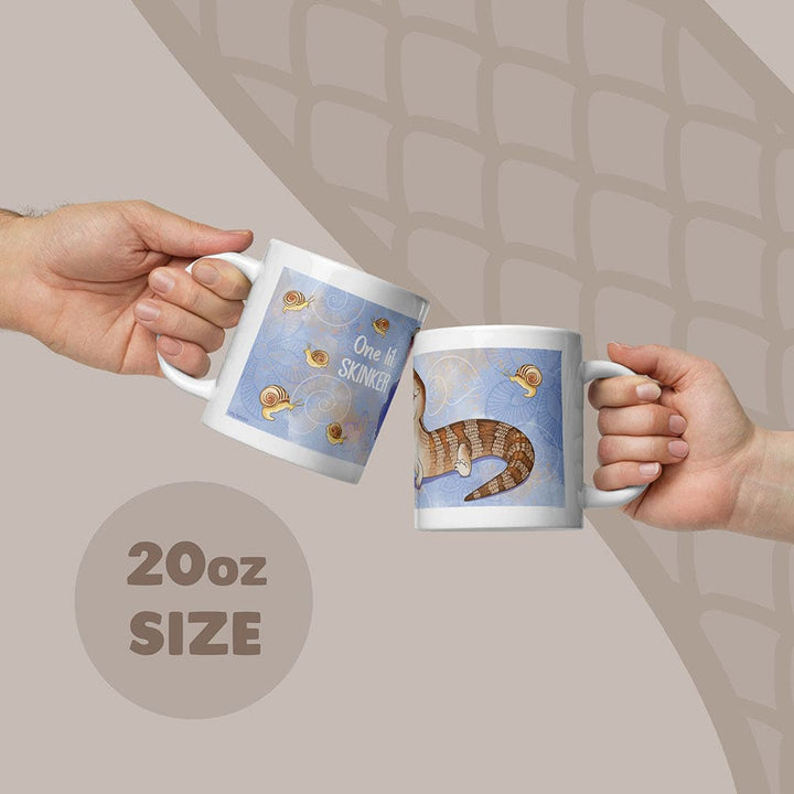 Two white ceramic mugs with an 20oz capacity, featuring a blue background with a brown and tan skink lizard illustration, small snail graphics, and the text One lil SKINKER printed in white on the side of the mugs.