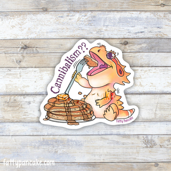 A die-cut sticker featuring a cartoon orange bearded dragon eating pancakes with a fork, placed on a whitewashed wood background. The sticker includes the text &