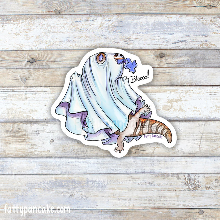 A die-cut sticker featuring a cartoon blue tongue skink, wearing a white sheet to look like a ghost, poking it&