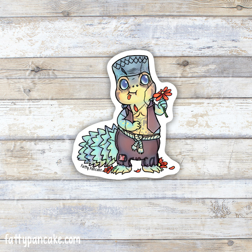 A die-cut sticker featuring a cartoon uromastyx lizard dressed up as Frankenstein wearing a brown vest and patchwork pants, holding a red flower, displayed on a light wood plank background.