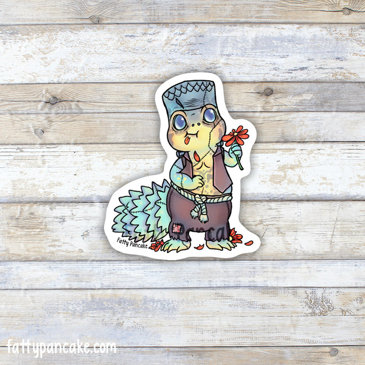 A die-cut sticker featuring a cartoon uromastyx lizard dressed up as Frankenstein wearing a brown vest and patchwork pants, holding a red flower, displayed on a light wood plank background.