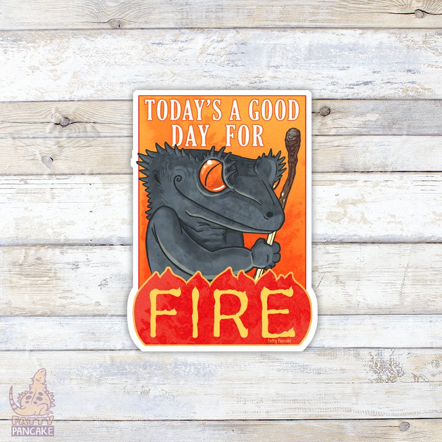 Cartoon grey crested gecko with a mischievous smile, holding a burnt match, holding a sword with 'Today's a good day for fire' text on an orange background, on a wooden surface.