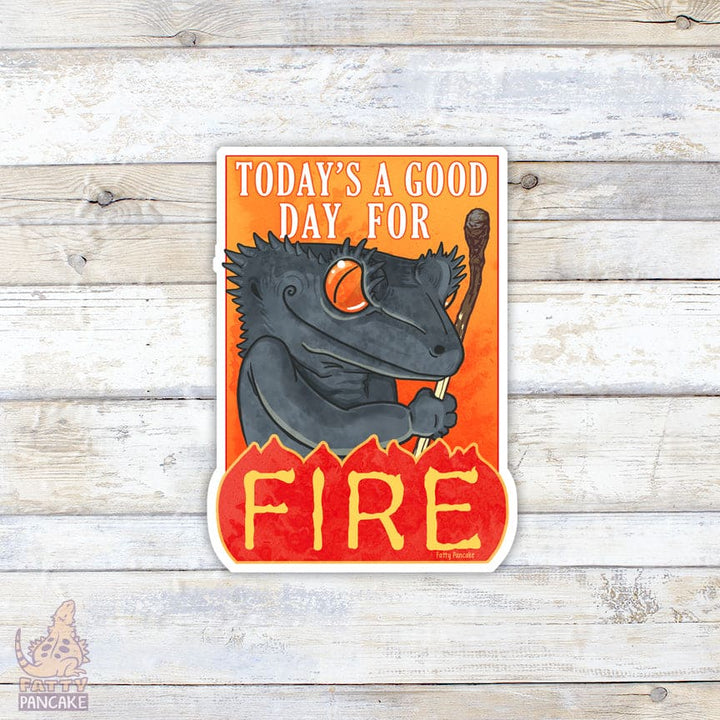 Cartoon grey crested gecko with a mischievous smile, holding a burnt match, holding a sword with 'Today's a good day for fire' text on an orange background, on a wooden surface.