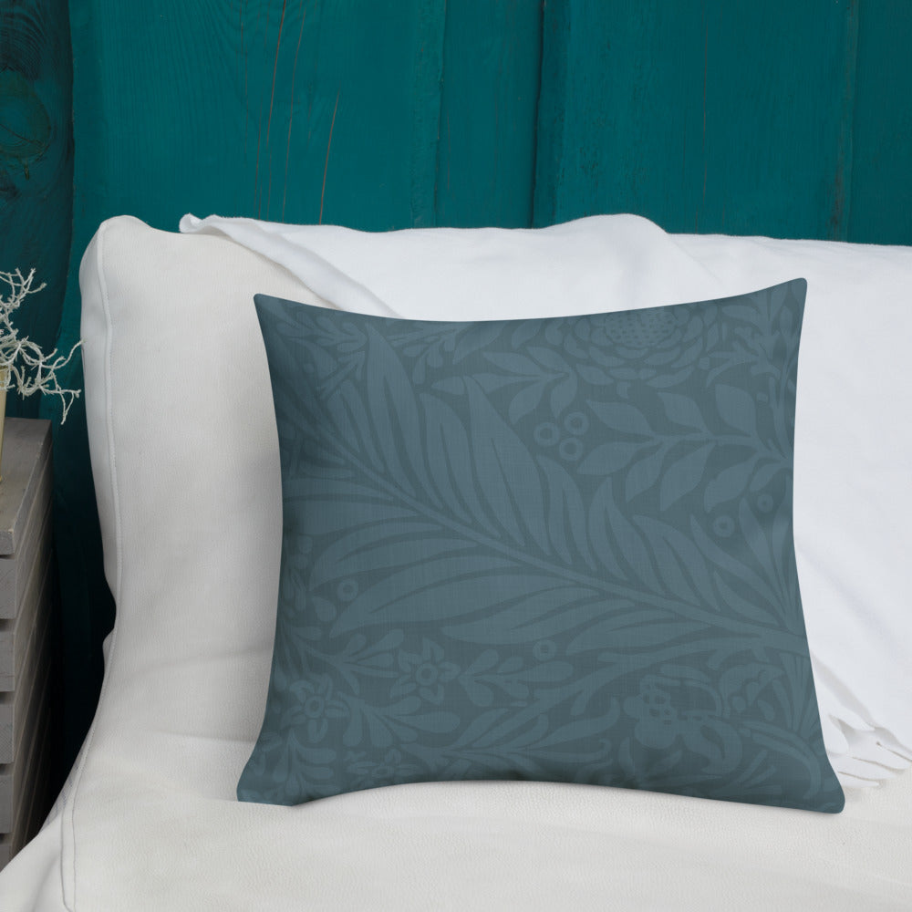 A square throw pillow in a muted slate blue-gray color with a subtle tonal botanical leaf pattern, resting on a white sofa in front of a teal wooden wall.