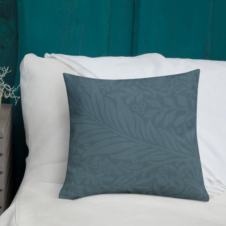 A square throw pillow in a muted slate blue-gray color with a subtle tonal botanical leaf pattern, resting on a white sofa in front of a teal wooden wall.
