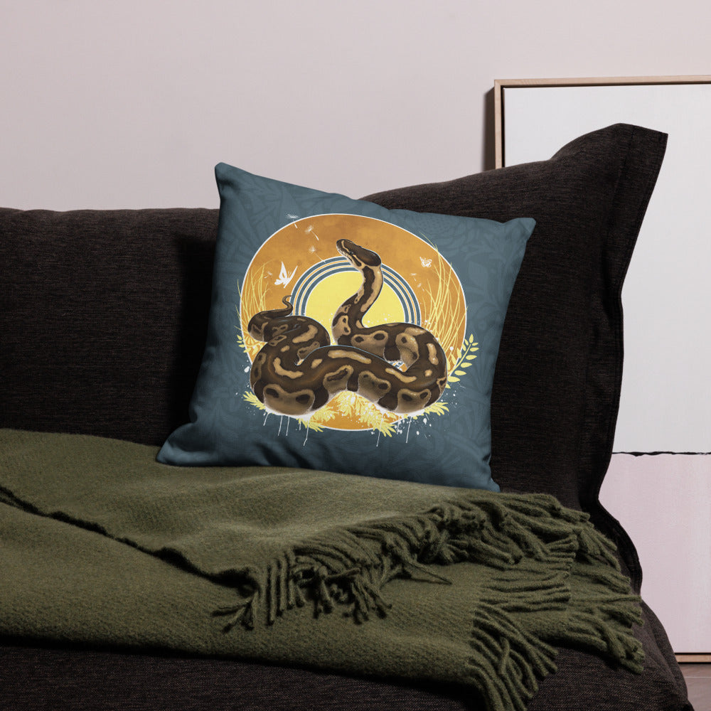 A square decorative throw pillow with a ball python snake illustration, styled on a dark sofa with a green throw blanket.