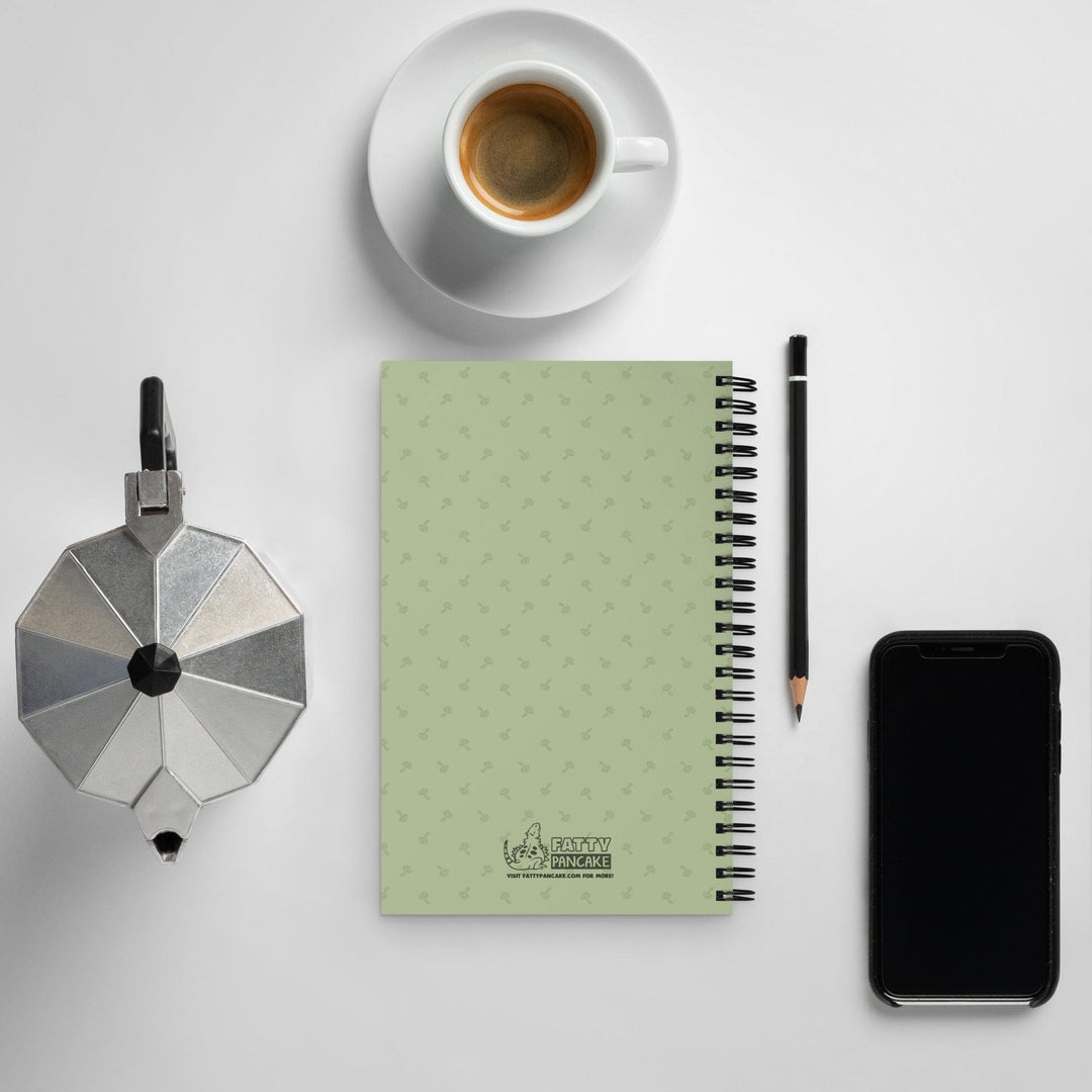 Rear depiction of a green journal with a mushroom pattern resting on a white table top and a cell phone, pencil, and coffee mug next to the notebook.