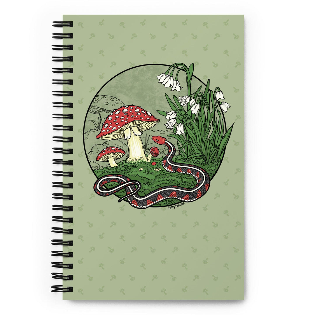 A green journal sitting on a bright white surface. The spiral notebook has a large graphic of a red sided garter snake with white flowers and red capped mushrooms. The style is line art with simple colors.