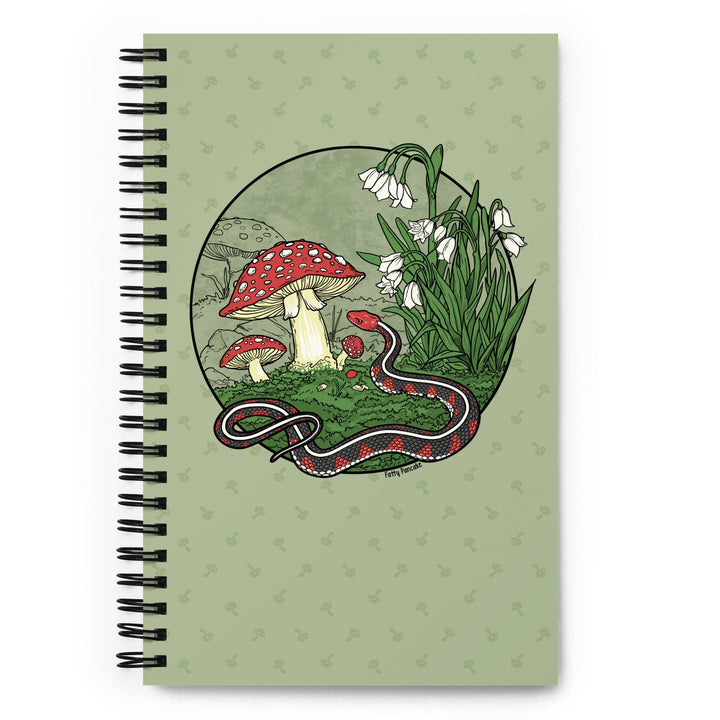 A green journal sitting on a bright white surface. The spiral notebook has a large graphic of a red sided garter snake with white flowers and red capped mushrooms. The style is line art with simple colors.