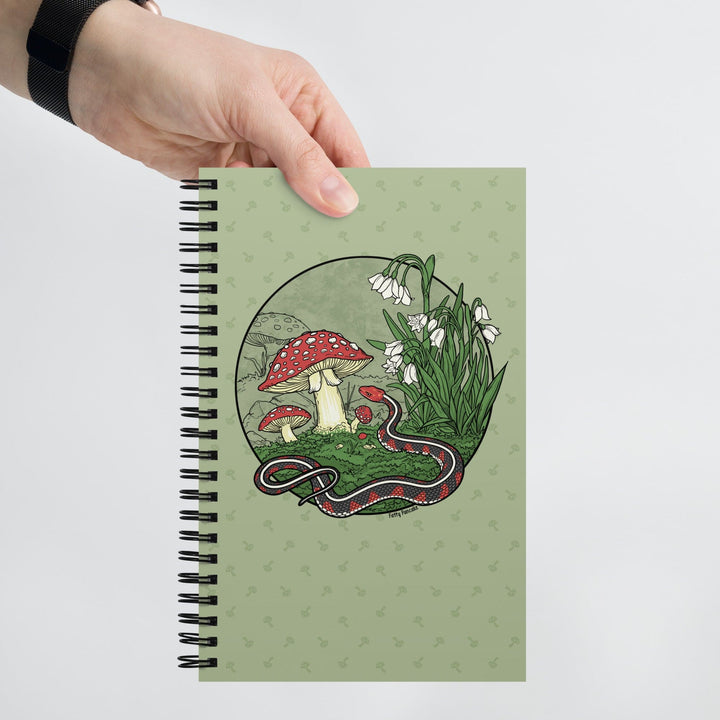 A green journal held by a hand against a white background. The spiral notebook has a large graphic of a red sided garter snake with white flowers and red capped mushrooms. The style is line art with simple colors.