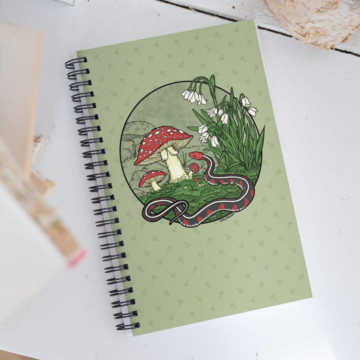 A green journal sitting on a white wooden surface. The spiral notebook has a large graphic of a red sided garter snake with white flowers and red capped mushrooms. The style is line art with simple colors.