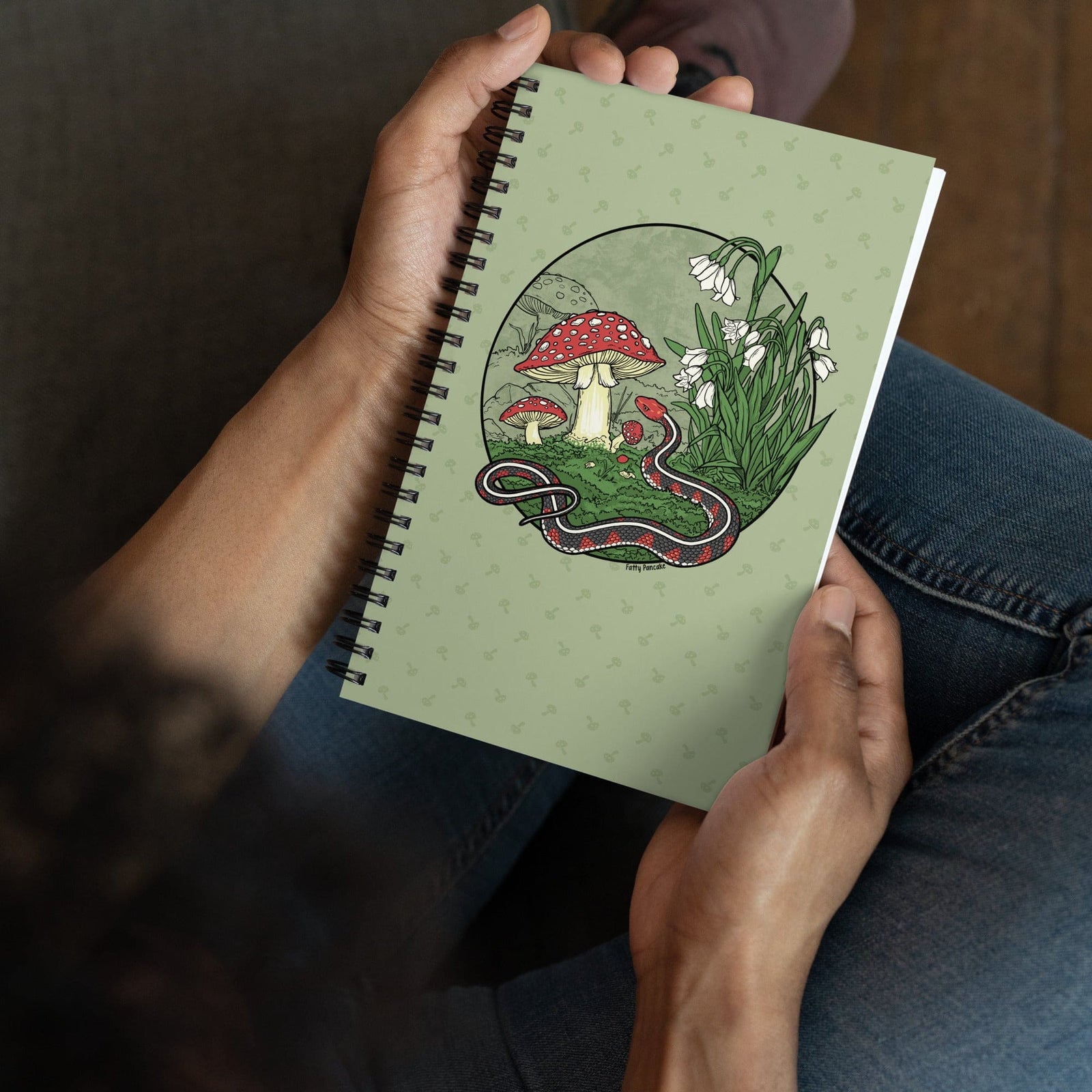 A green journal held in a persona's hands. The spiral notebook has a large graphic of a red sided garter snake with white flowers and red capped mushrooms. The style is line art with simple colors.