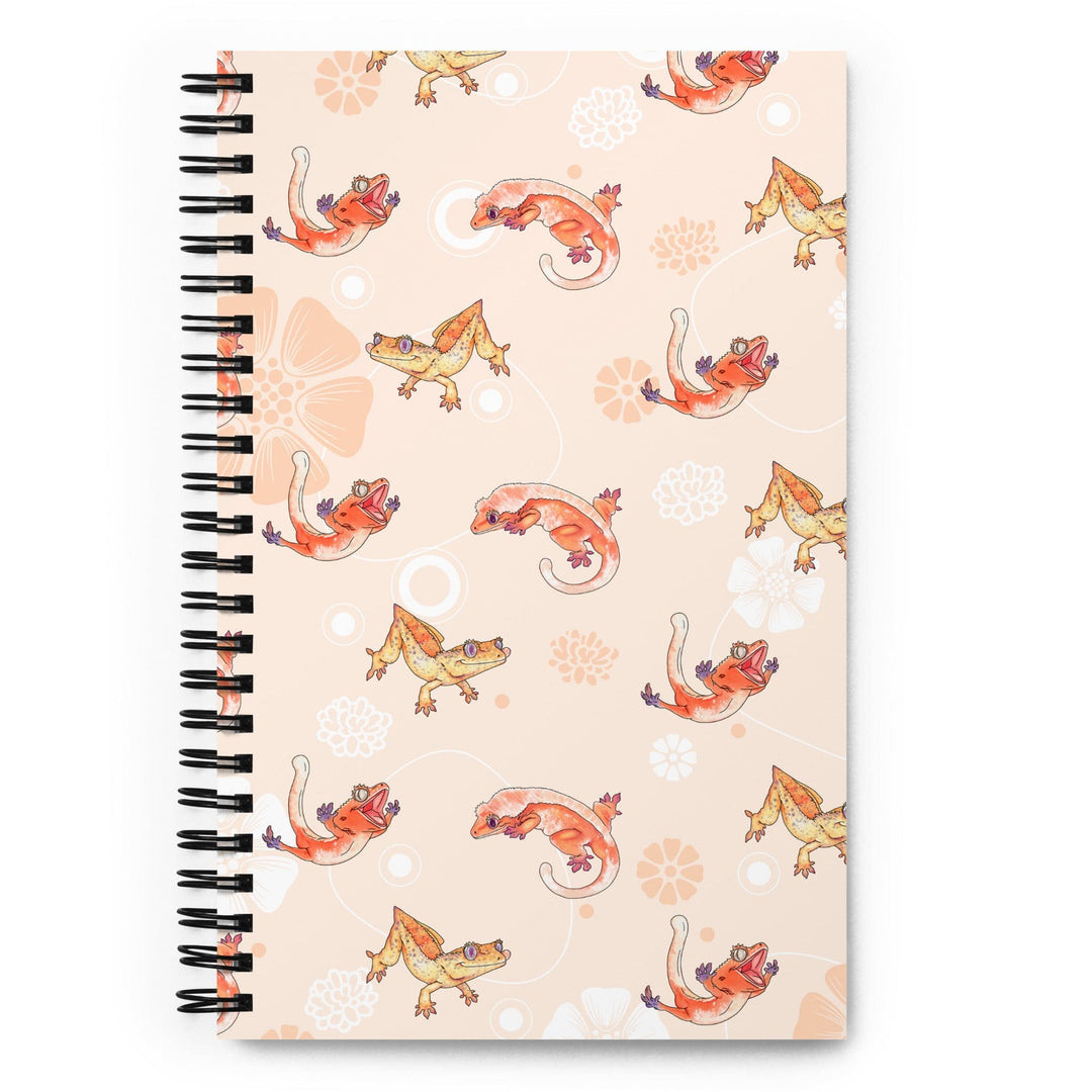 A peach colored journal with a flower pattern and many cartoon crested geckos sprinkled in the pattern, the journal rests on a blank white background. This is the front of the journal.