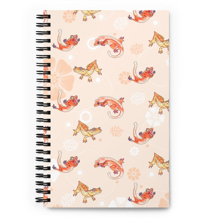 A peach colored journal with a flower pattern and many cartoon crested geckos sprinkled in the pattern, the journal rests on a blank white background. This is the front of the journal.