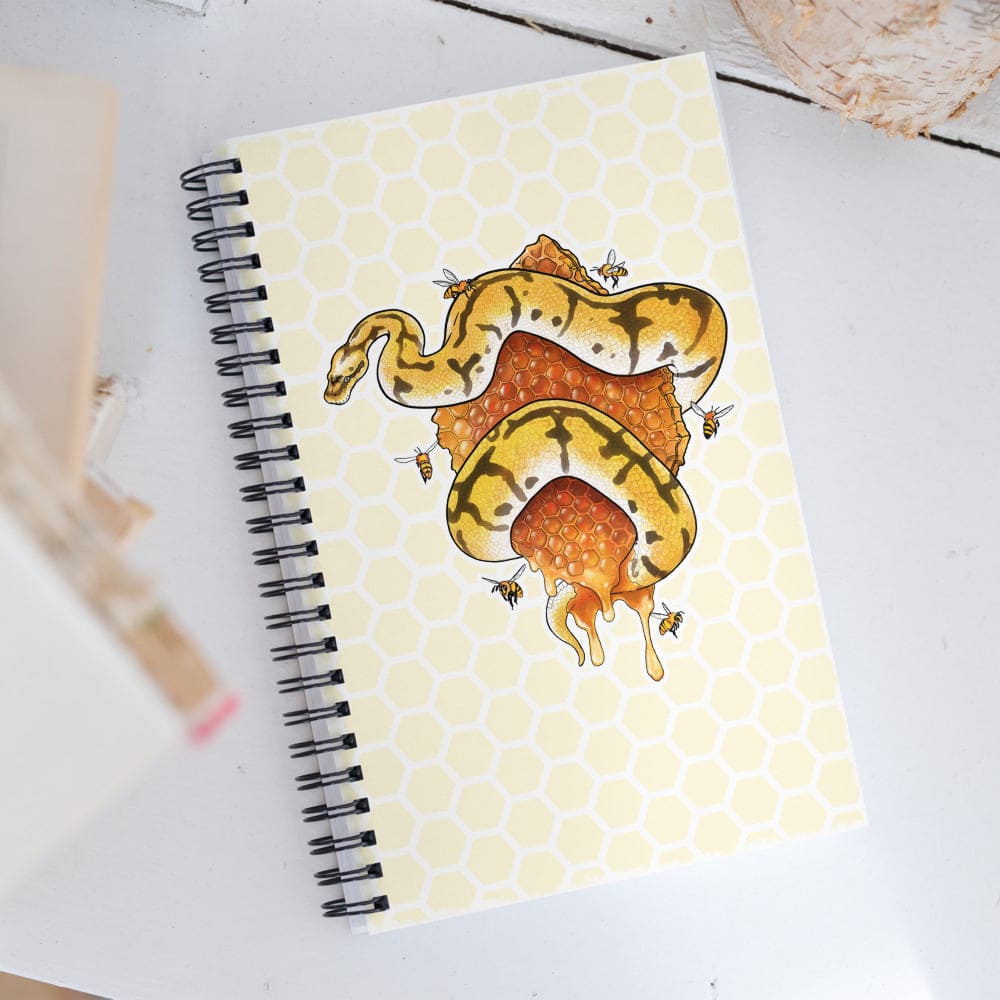 A spiral-bound notebook with a pale yellow honeycomb-patterned cover, featuring an illustration of a yellow and black snake wrapped in orange honeycomb with bees, photographed on a white surface.
