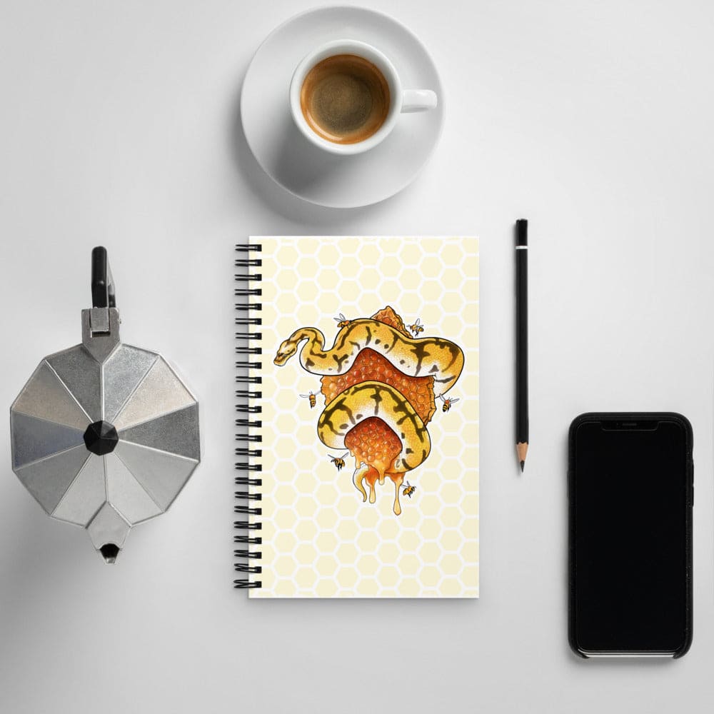 A spiral-bound notebook with a yellow and orange snake illustration on a light honeycomb-patterned cover, styled in a flat lay with a cup of coffee, a moka pot, a pencil, and a smartphone on a light gray background.