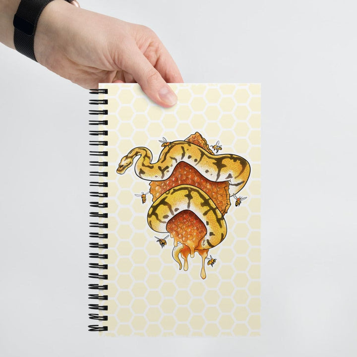 A hand holds a spiral-bound notebook against a light gray background. The notebook cover has a pale yellow honeycomb pattern and features an illustration of a yellow and black snake wrapped around a honeycomb with dripping honey.