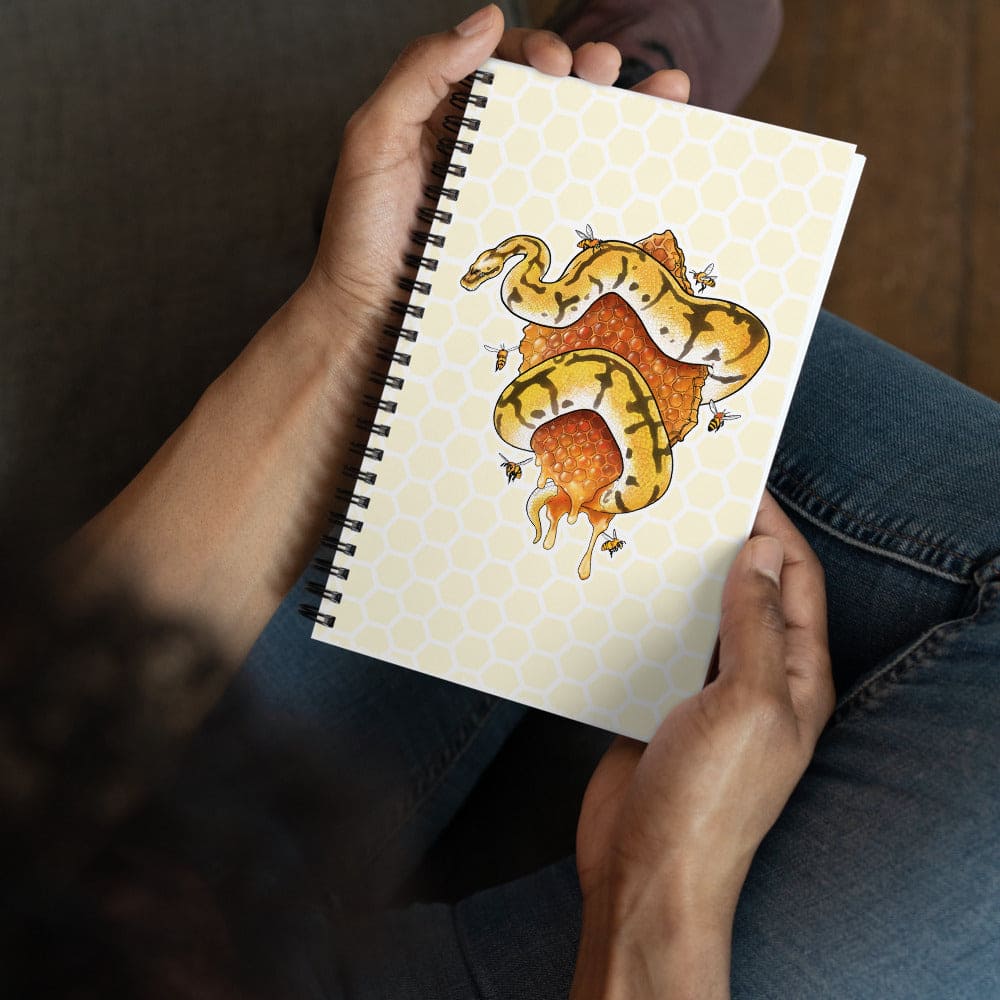A person holding a spiral-bound notebook with a yellow and orange snake coiled around a piece of honeycomb with bees  on the cover, shown from above while seated wearing blue jeans.