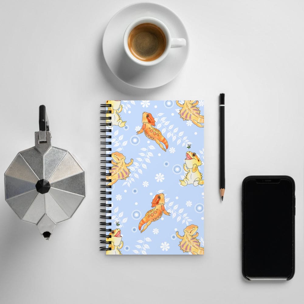 A spiral-bound notebook with a light blue cover featuring a pattern of cartoon bearded dragon lizards and white flowers, styled in a flat-lay arrangement on a light gray surface alongside a cup of coffee, a moka pot, a pencil, and a smartphone.