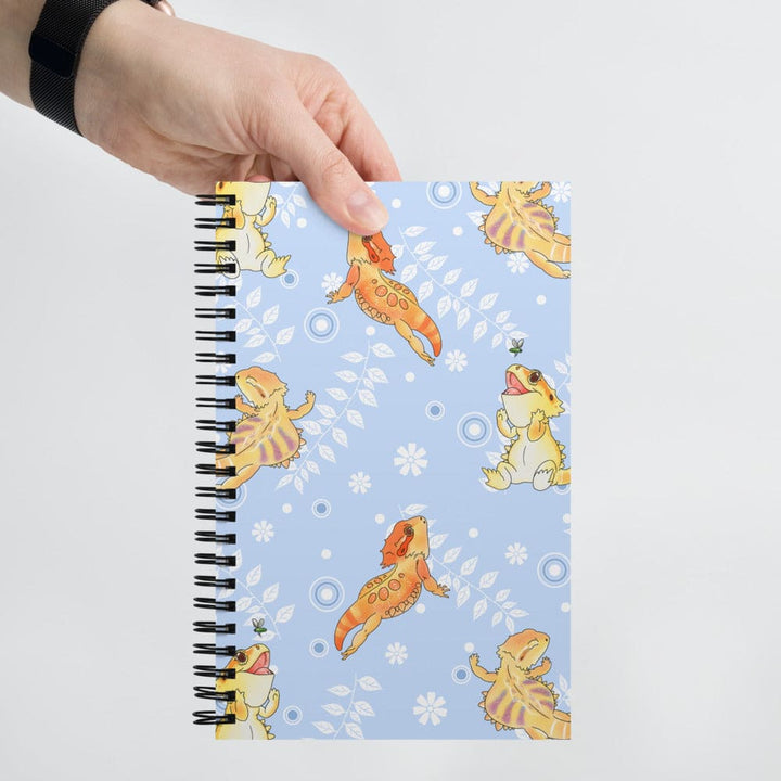 A hand holds a spiral-bound notebook with a light blue cover decorated with cartoon bearded dragon lizards  in orange and yellow, and white leaves on a plain light gray background.