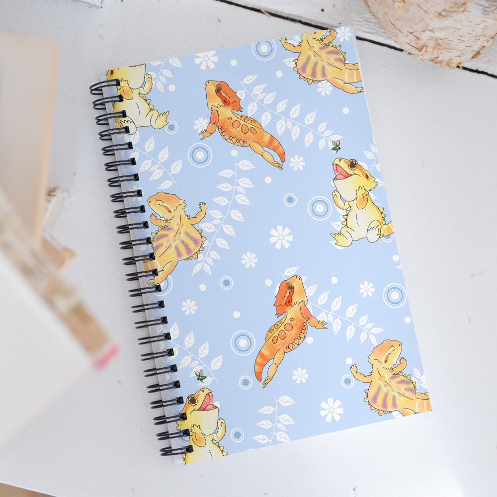 A spiral-bound notebook with a light blue cover decorated with cartoon bearded dragon lizards in yellow and orange, and white leaves and circles, photographed on a white tabletop.