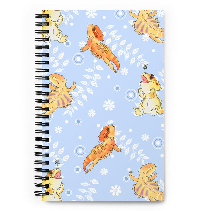 A spiral-bound notebook with a light blue cover decorated with cartoon bearded dragons and white floral motifs, shown on a plain white background.
