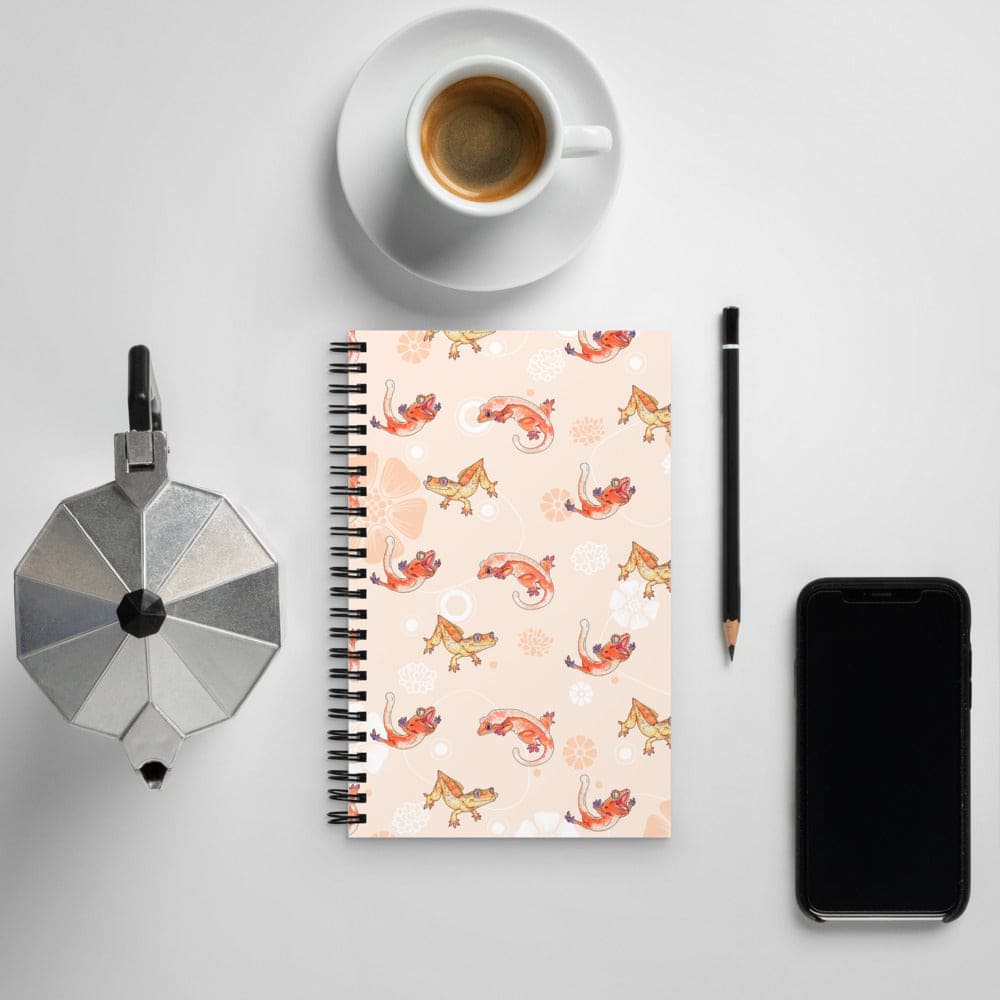 Notebook with a pink and peach pattern with cartoon crested geckos, a coffee cup, phone, and other items on a light gray background