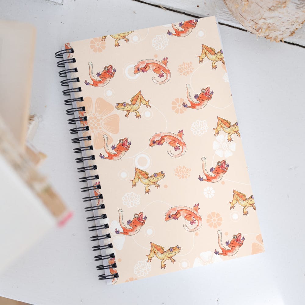 A spiral-bound notebook with a beige cover decorated with a repeating pattern of orange and yellow crested geckos, photographed on a white tabletop.
