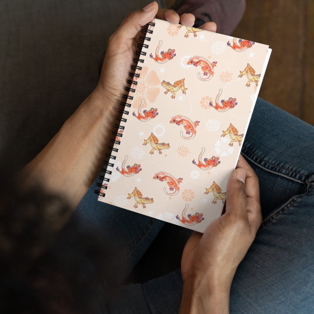 A person holding a spiral-bound notebook with a light peach cover decorated with a repeating pattern of orange and yellow crested geckos and small white floral motifs, shown from an overhead angle against blue jeans.