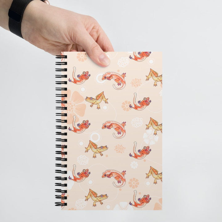 A hand holding a spiral-bound notebook with a peach cover decorated with a repeating pattern of orange and yellow crested geckos and white floral motifs, set against a plain light gray background.