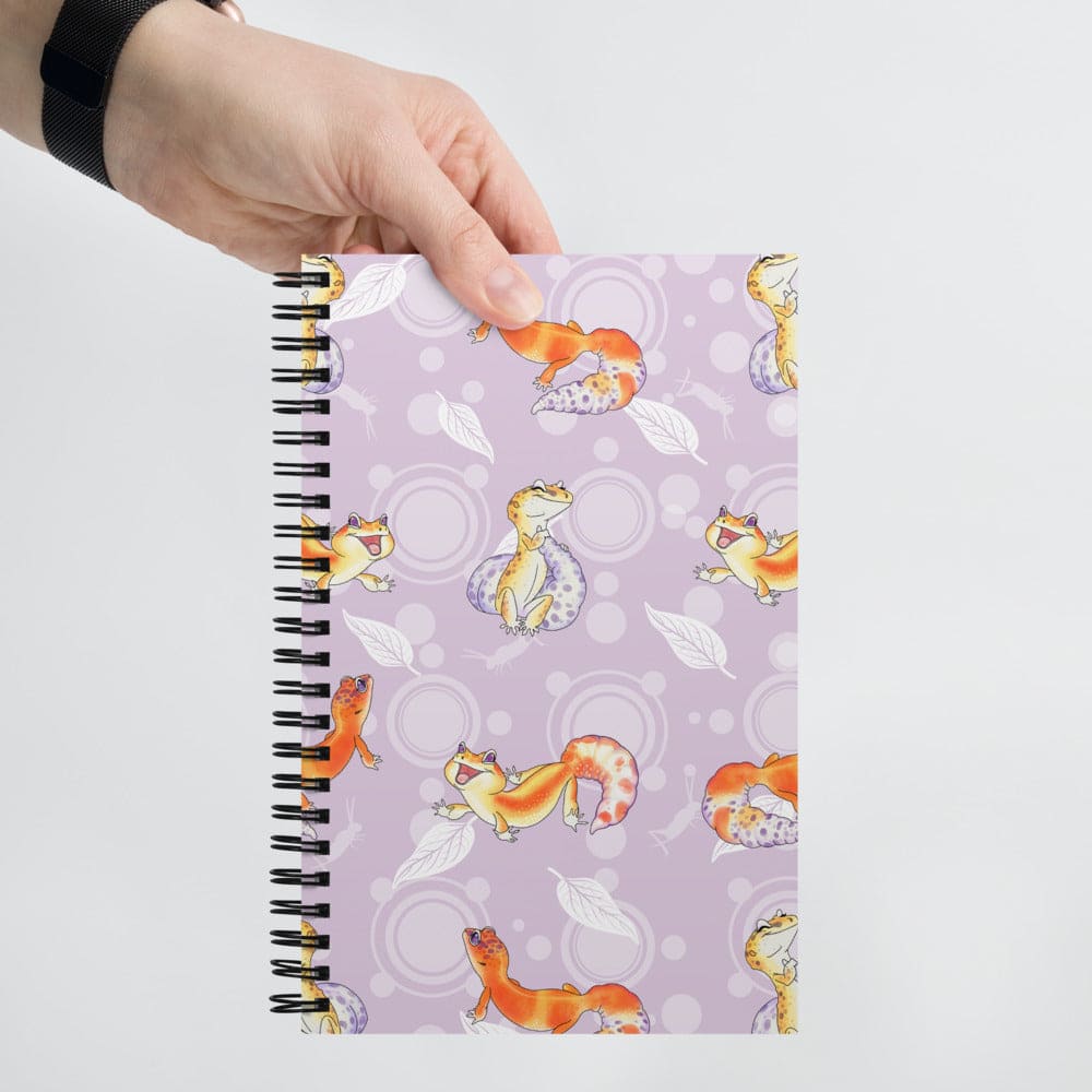 A hand holds a spiral-bound notebook with a lavender cover decorated with a repeating pattern of orange and yellow cartoon leopard geckos, white leaves, and circular motifs.