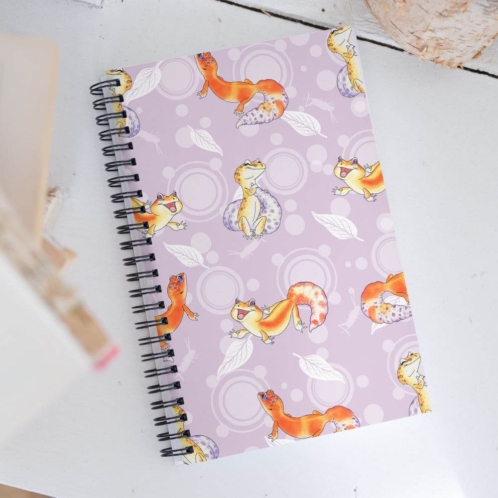 A spiral-bound notebook with a light purple cover featuring a playful pattern of cartoon orange and yellow leopard geckos, white leaves, and circular motifs, photographed on a white tabletop.