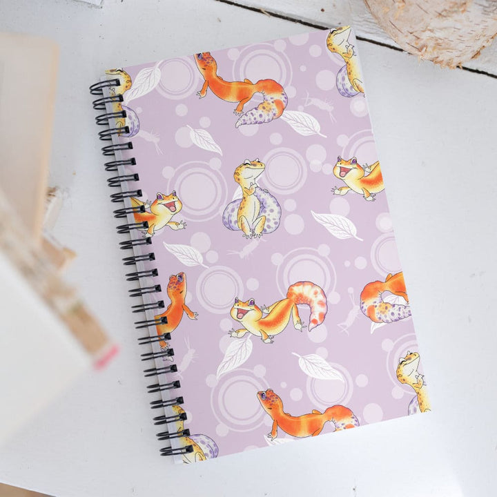 A spiral-bound notebook with a light purple cover featuring a playful pattern of cartoon orange and yellow leopard geckos, white leaves, and circular motifs, photographed on a white tabletop.