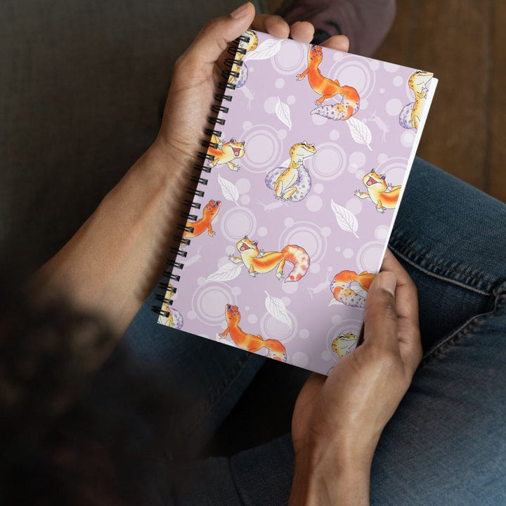 A person wearing blue jeans holds a spiral-bound notebook with a lavender cover decorated with orange leopard geckos and white leaf patterns.