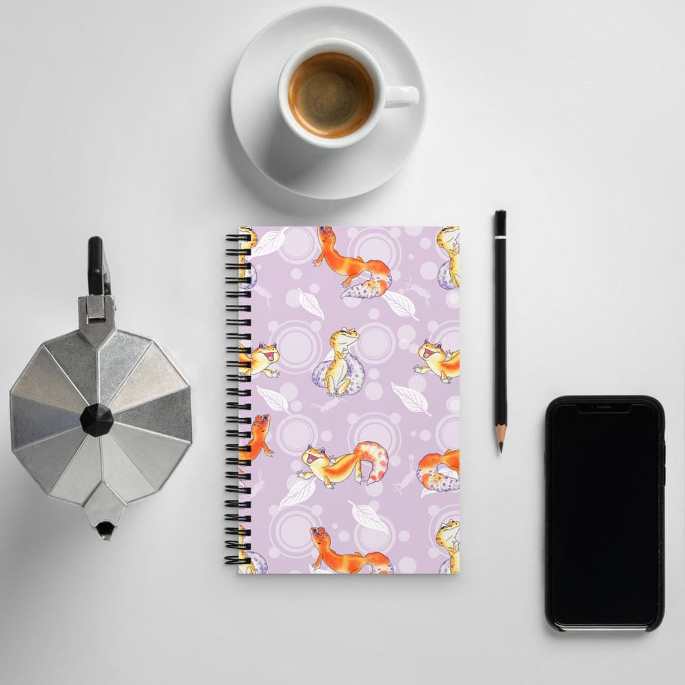 A spiral-bound notebook with a light purple cover featuring a pattern of orange and yellow leopard geckos, styled in a flat-lay arrangement with a cup of coffee, a moka pot, a pencil, and a smartphone on a light gray background.