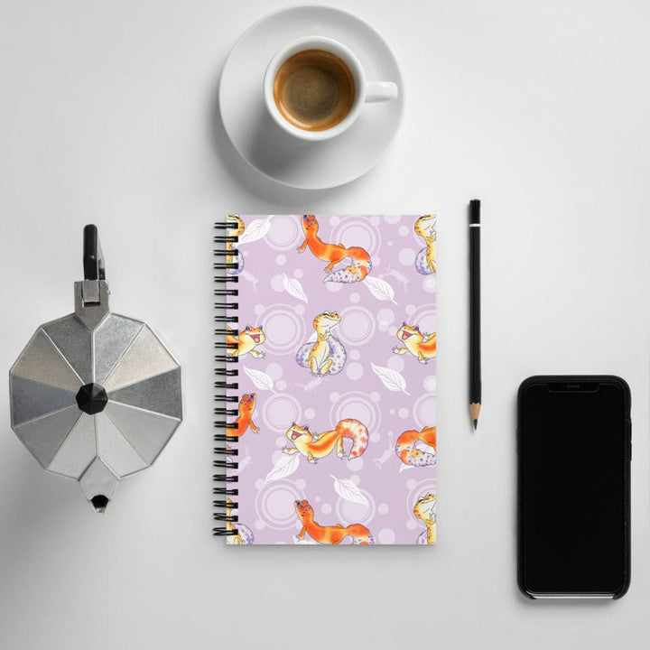 A spiral-bound notebook with a light purple cover featuring a pattern of orange and yellow leopard geckos, styled in a flat-lay arrangement with a cup of coffee, a moka pot, a pencil, and a smartphone on a light gray background.