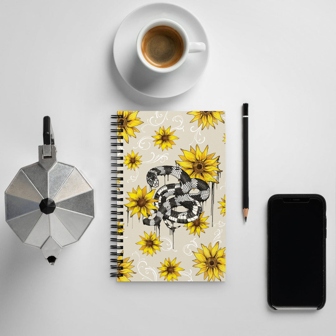 A spiral-bound notebook with a yellow sunflower and black-and-white snake illustration, styled in a flat lay on a light gray surface with a cup of coffee, a moka pot, a pencil, and a smartphone.
