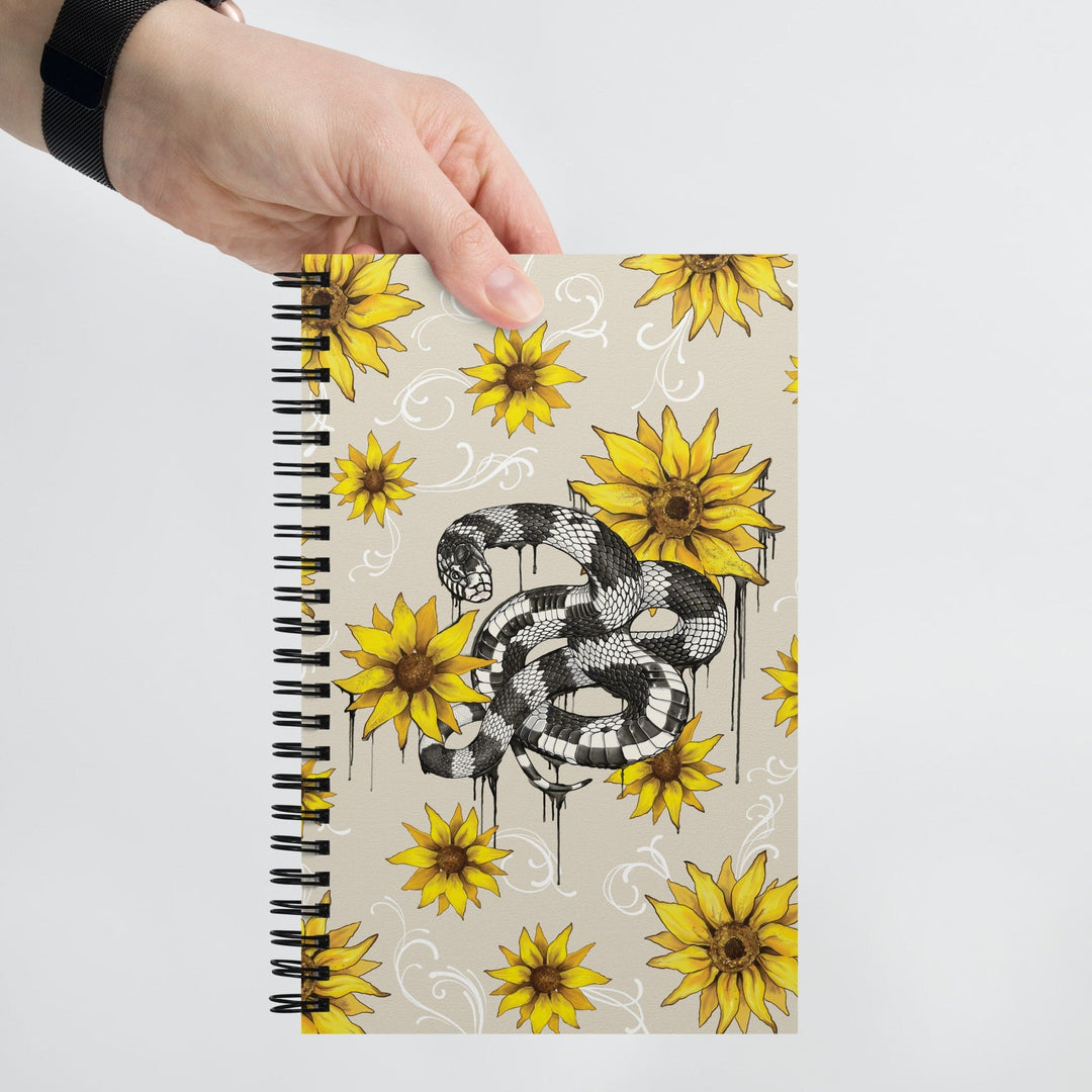 A hand holds a spiral-bound notebook featuring a black and white snake illustration surrounded by yellow sunflowers on a light beige background.