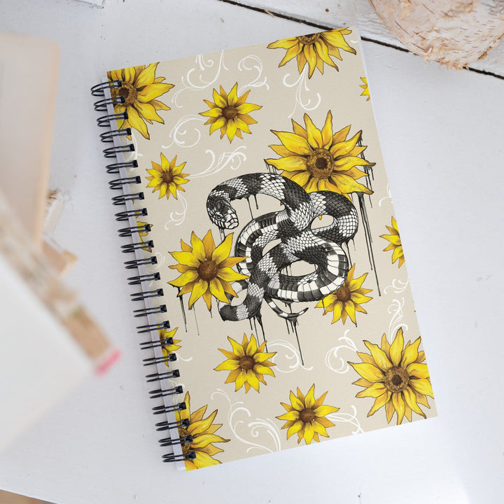 A spiral-bound notebook with a beige cover featuring a black and white snake illustration and yellow sunflower designs, photographed on a white tabletop.