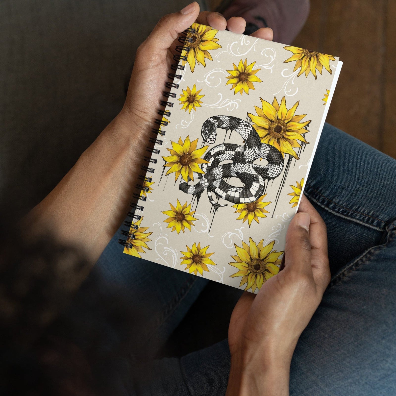 A person holding a spiral-bound notebook with a beige cover decorated with yellow sunflowers and a black-and-white snake illustration, shown from above against a casual indoor background.
