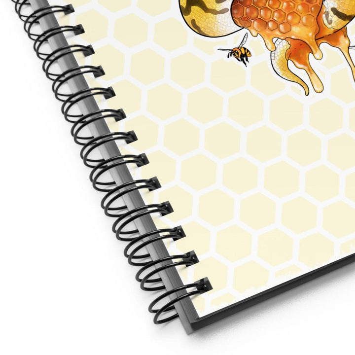 A close-up of a spiral-bound notebook showing the cover design with a pale yellow honeycomb pattern, dripping orange honey, a bee, and part of a floral illustration. The notebook has a black spiral binding and is photographed on a white background.
