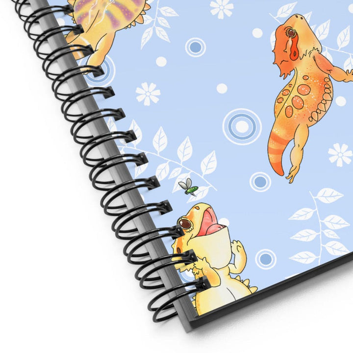 A spiral-bound notebook shown at an angle on a white background, featuring a light blue cover decorated with cartoon bearded dragons, white leaves, and bubbles.