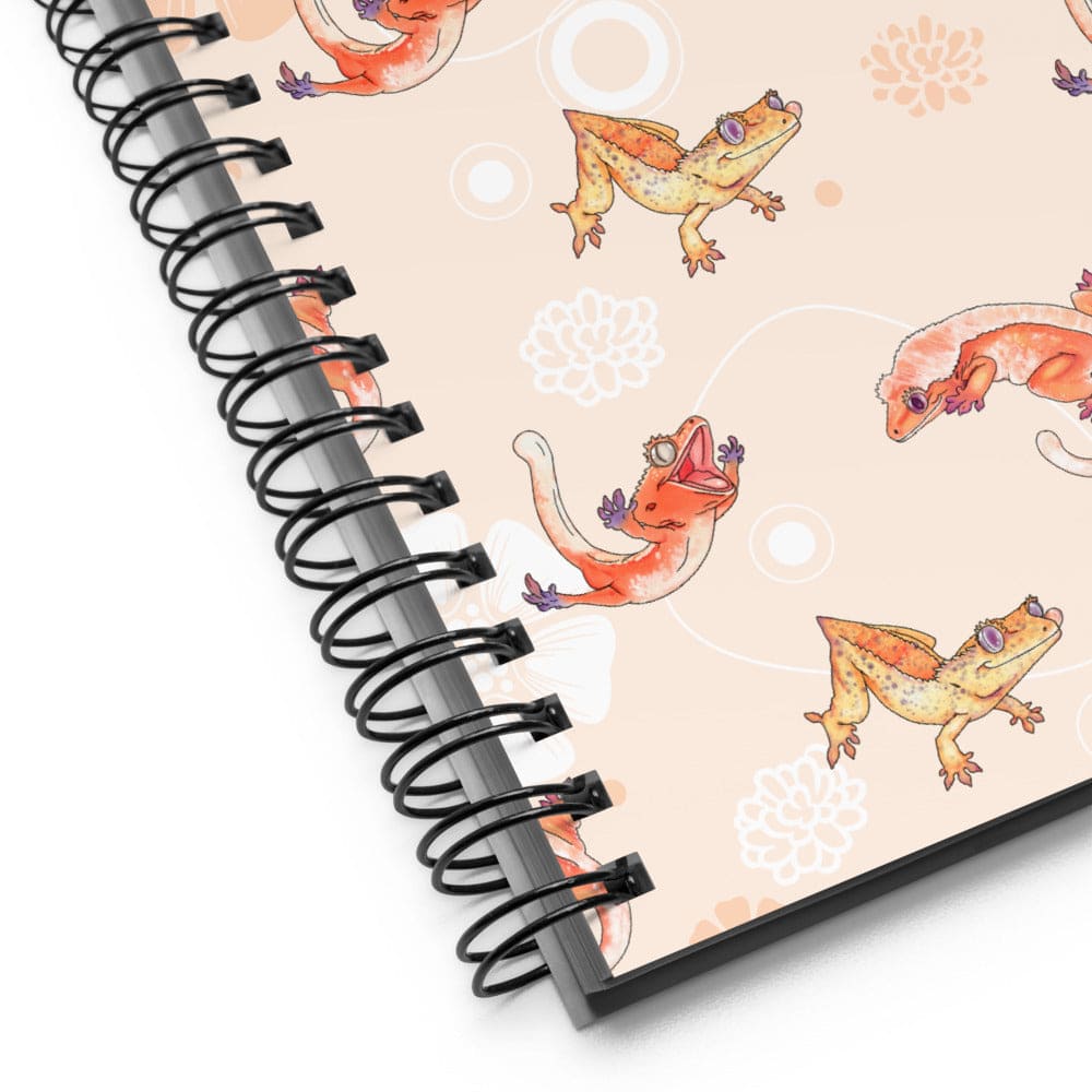 A spiral-bound notebook shown at an angle on a white background. The notebook cover has a light peach background with a repeating pattern of orange crested geckos and white floral motifs, bound with a black metal spiral.