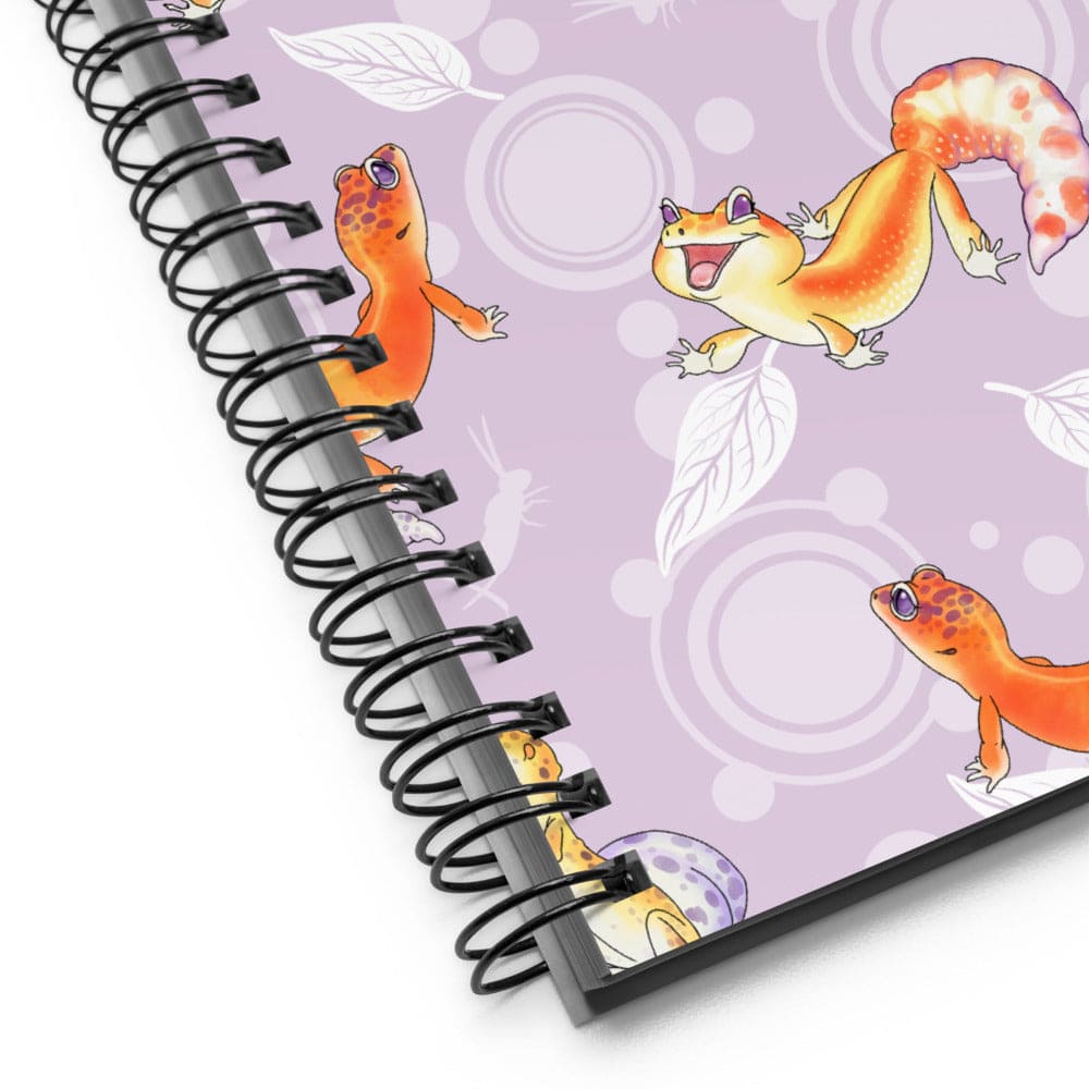 A spiral-bound notebook shown at an angle on a white background. The notebook cover is light purple with cartoon gecko illustrations in orange, yellow, and white, along with white leaf and circular patterns.